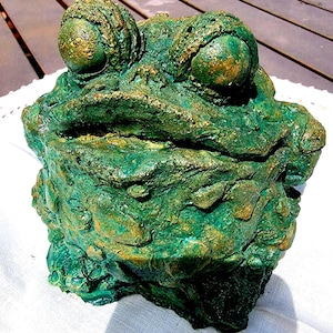 May include: A detailed, textured sculpture of a frog with large, bulbous eyes. The frog is primarily green with gold accents, giving it a weathered appearance. The sculpture is likely made of a durable material.