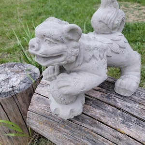 May include: A gray concrete Foo Dog statue, a traditional Chinese guardian figure. The statue is in a crouching position, holding a ball, with detailed features and a textured surface. It is placed on a weathered wooden log, with green grass in the background.