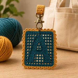 Puede incluir: Un llavero rectangular de ganchillo con una letra A de color verde azulado oscuro centrada en un patrón de cuadrícula, bordeado por un borde festoneado de color amarillo mostaza. La pieza está unida a un lazo de ganchillo amarillo mostaza y un mosquetón de metal color bronce.