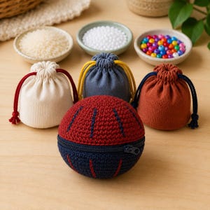 Può includere: Una pochette sferica all'uncinetto bicolore rossa e blu navy con cerniera, posizionata davanti a tre piccoli sacchetti di stoffa con coulisse nei colori crema, grigio e ruggine, dotati di cordini a contrasto con nodi.