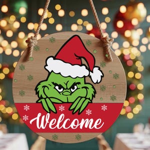 May include: Round wooden Christmas sign featuring the Grinch wearing a Santa hat, with the word "Welcome" in white script on a red banner. The sign has a light brown background with snowflake and green dot accents, and is suspended by a natural rope.