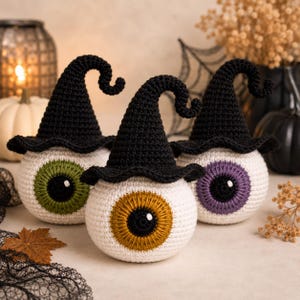 May include: Three spherical crochet eyeballs, each wearing a pointed black witch hat with a curled tip. The eyeballs have white sclera and irises in green, gold, and purple, each featuring a black pupil with a small white highlight stitch.