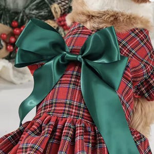 May include: A red plaid dog dress with a large green satin bow. The dress features a classic tartan pattern with black, white, and yellow accents. The bow is a vibrant green, adding a festive touch to the garment. The dress has a faux fur collar.