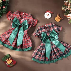 May include: Two plaid dog dresses in red, black, and green, each with a large green satin bow. The dresses have a pleated skirt and green trim. Christmas decorations surround the dresses.