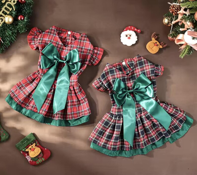 May include: Two plaid dog dresses in red, black, and green, each with a large green satin bow. The dresses have a pleated skirt and green trim. Christmas decorations surround the dresses.