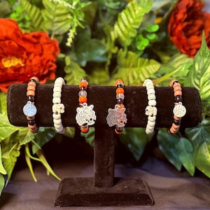 May include: Six beaded bracelets displayed on a black stand. The bracelets feature various designs, including skulls, tiger faces, and round silver charms. The beads are in colors like white, orange, black, and brown. The background includes red flowers and green foliage.
