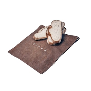 Può includere: Un sacchetto per il pane in lino marrone con la parola "bread" ricamata in bianco. Diverse fette di pane poggiano sopra il sacchetto, mostrando l'uso previsto del prodotto. Il sacchetto è rettangolare e ha un design semplice e minimalista.