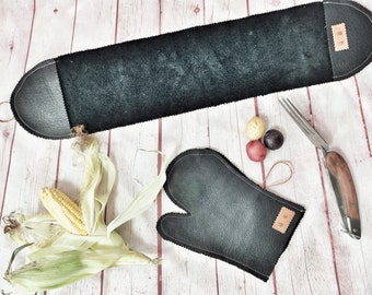 May include: Black leather oven mitt and a long, rectangular black leather hot pad with white stitching. A corn cob, potatoes, and a knife with a wooden handle are also present. The items are arranged on a light wood surface.