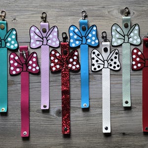 May include: A collection of eight colorful wristlet keychains. Each keychain features a bow design with polka dots or glitter, attached to a matching strap. Colors include teal, pink, purple, red, blue, white, and green. The keychains have a metal clasp.