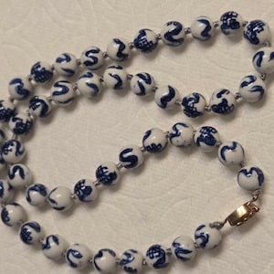 May include: A necklace composed of round white ceramic beads, each featuring a hand-painted blue pattern. The beads are strung together on a cord and secured with a gold-toned metal clasp. The design consists of repeating blue motifs on a smooth, glossy white surface.