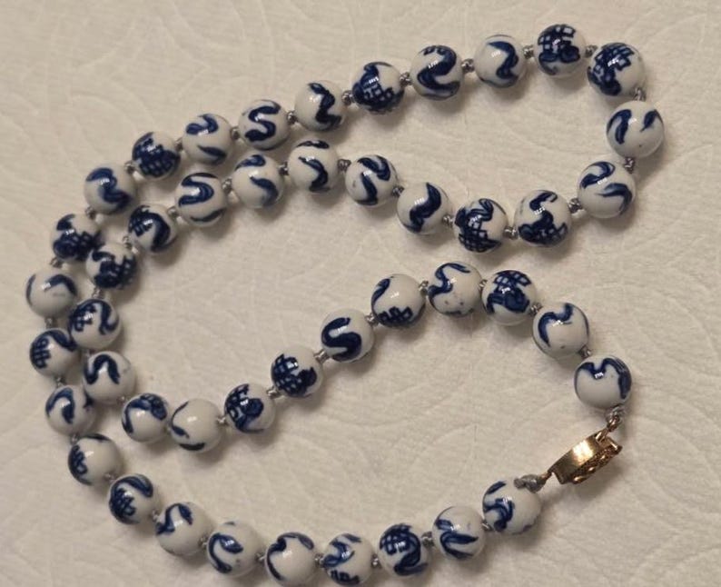 May include: A necklace composed of round white ceramic beads, each featuring a hand-painted blue pattern. The beads are strung together on a cord and secured with a gold-toned metal clasp. The design consists of repeating blue motifs on a smooth, glossy white surface.