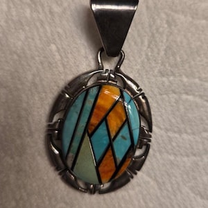 May include: An oval pendant featuring a geometric inlay of turquoise, orange, and pale green stone segments separated by thin black lines. The inlay is set within a polished silver frame with a decorative notched edge and a triangular bail at the top.