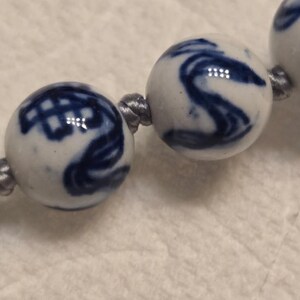 May include: A string of spherical ceramic beads featuring a white base with hand-painted blue patterns. The beads are connected by a thin, grey cord, with each bead displaying unique, swirling, and geometric blue ink designs across the surface.
