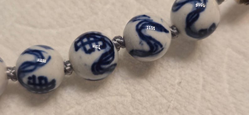 May include: A string of spherical ceramic beads featuring a white base with hand-painted blue patterns. The beads are connected by a thin, grey cord, with each bead displaying unique, swirling, and geometric blue ink designs across the surface.