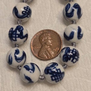 May include: A necklace featuring round white ceramic beads with blue hand-painted patterns, strung on a grey cord. A 1961 copper penny is placed between the beads to provide a scale reference for the size of the individual spherical components.
