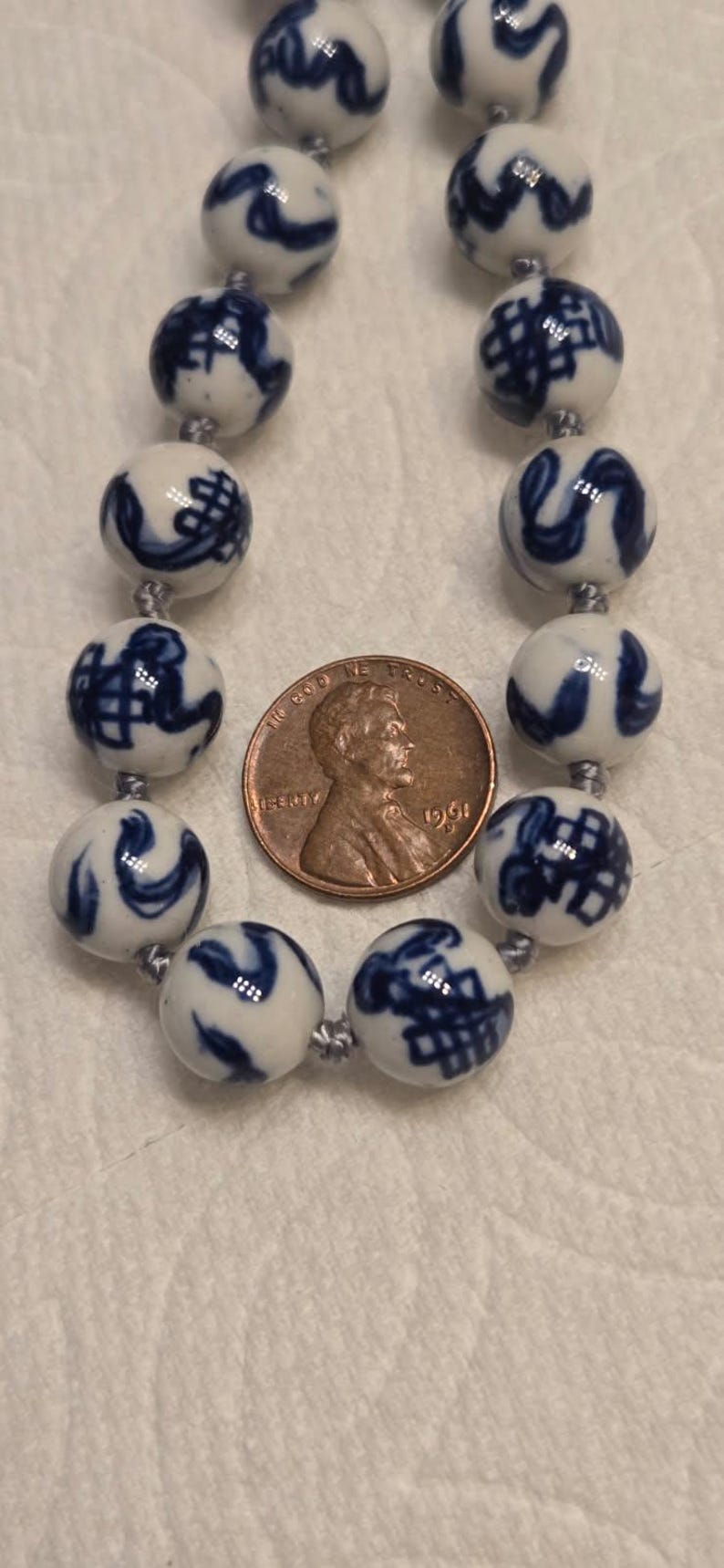 May include: A necklace featuring round white ceramic beads with blue hand-painted patterns, strung on a grey cord. A 1961 copper penny is placed between the beads to provide a scale reference for the size of the individual spherical components.