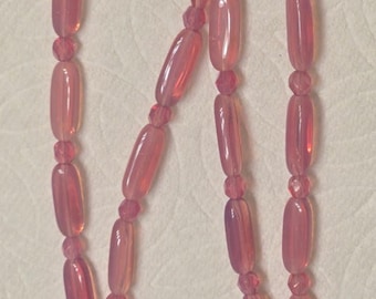 May include: A delicate pink necklace featuring elongated, oval-shaped beads interspersed with smaller, round beads. The necklace has a gold-colored clasp. The beads have a translucent quality, creating a soft, glowing appearance. This jewelry piece is suitable for various occasions.