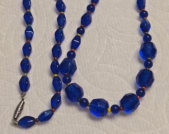 May include: A vintage necklace featuring faceted, deep blue glass beads. The necklace has a variety of bead shapes and sizes, with small yellow and red accent beads. It is secured with a silver-tone clasp.