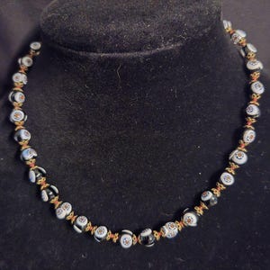 May include: A necklace featuring black and white glass beads with a floral pattern, incorporating blue and orange details. The beads are strung on a red cord, connected by gold-toned elements. The necklace is displayed on a black velvet bust.