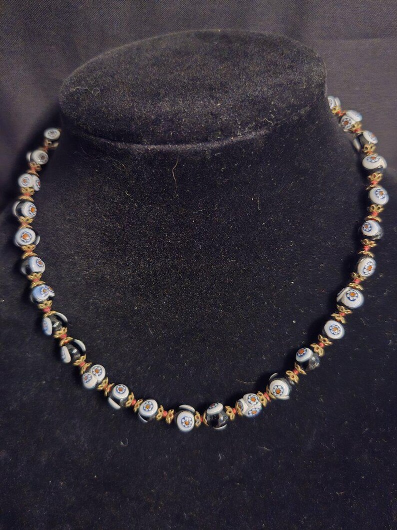 May include: A necklace featuring black and white glass beads with a floral pattern, incorporating blue and orange details. The beads are strung on a red cord, connected by gold-toned elements. The necklace is displayed on a black velvet bust.