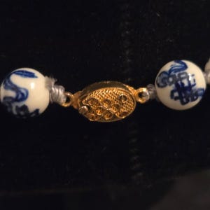 May include: A necklace featuring round white ceramic beads decorated with blue painted patterns. The beads are strung together and secured with an oval-shaped gold-toned metal clasp that has a textured, filigree-like surface design.