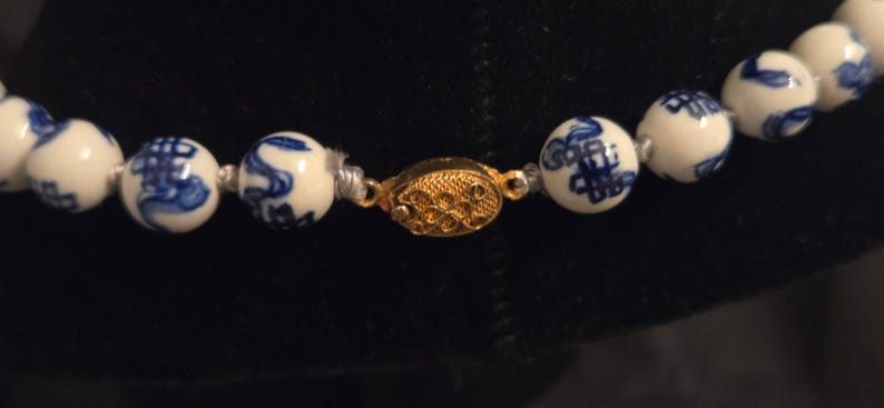 May include: A necklace featuring round white ceramic beads decorated with blue painted patterns. The beads are strung together and secured with an oval-shaped gold-toned metal clasp that has a textured, filigree-like surface design.