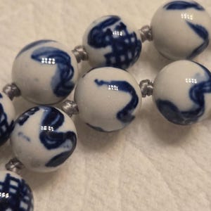 May include: A string of spherical white ceramic beads featuring hand-painted blue patterns. Each bead is connected by a thin grey cord, creating a continuous strand. The surface of the beads is smooth and glossy, displaying various abstract blue designs.