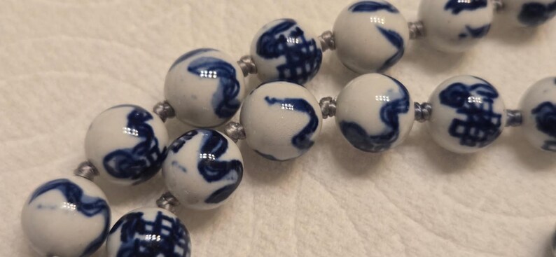 May include: A string of spherical white ceramic beads featuring hand-painted blue patterns. Each bead is connected by a thin grey cord, creating a continuous strand. The surface of the beads is smooth and glossy, displaying various abstract blue designs.