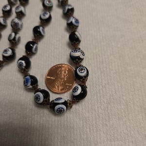 May include: A vintage necklace with black and white glass beads, each with a floral pattern. The beads are strung on a reddish-brown cord. A penny is next to the necklace for size comparison.