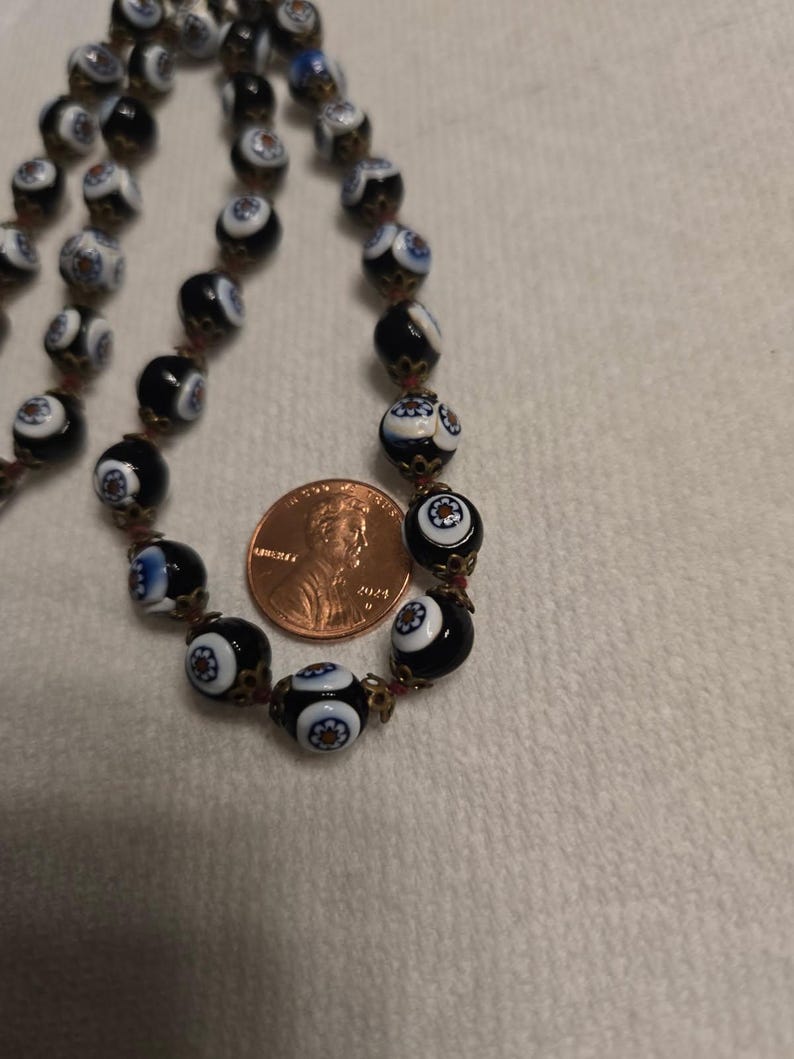 May include: A vintage necklace with black and white glass beads, each with a floral pattern. The beads are strung on a reddish-brown cord. A penny is next to the necklace for size comparison.