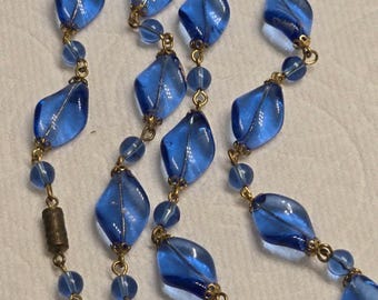 May include: A vintage necklace featuring translucent blue glass beads in a teardrop shape, strung together with small round beads and gold-toned metal chain. The necklace has a classic design.