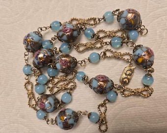 May include: A necklace featuring a gold-tone chain interspersed with round, light blue glass beads and larger, decorative beads painted with floral patterns in pink, yellow, and blue, accented with metallic gold leaf detailing throughout.