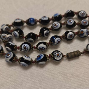 May include: Close-up of a beaded necklace with black, white, and blue glass beads. The beads have a floral design and are strung on a red cord. The necklace has a metal clasp.