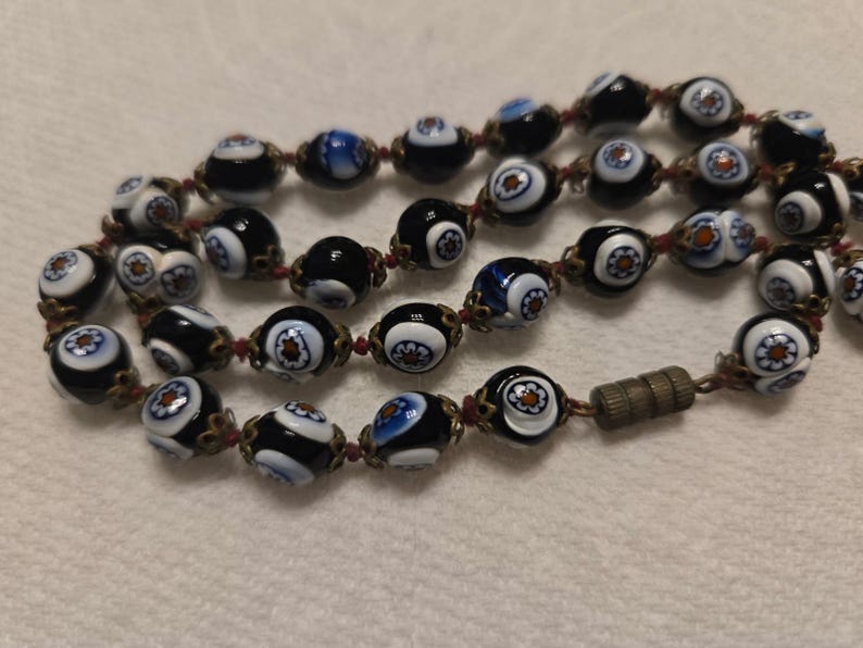 May include: Close-up of a beaded necklace with black, white, and blue glass beads. The beads have a floral design and are strung on a red cord. The necklace has a metal clasp.