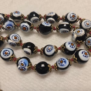 May include: A necklace with black and white glass beads, each decorated with a floral pattern. The beads are linked with red string and gold-coloured metal. A vintage-style necklace.