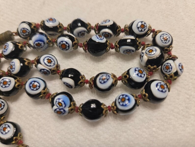 May include: A necklace with black and white glass beads, each decorated with a floral pattern. The beads are linked with red string and gold-coloured metal. A vintage-style necklace.