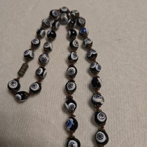 May include: A vintage necklace with patterned beads in black, white, and blue. The beads have a floral design and are strung together with a dark red cord. The necklace has a metal clasp.
