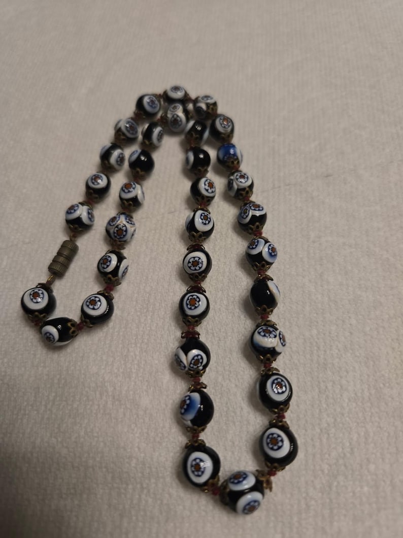 May include: A vintage necklace with patterned beads in black, white, and blue. The beads have a floral design and are strung together with a dark red cord. The necklace has a metal clasp.