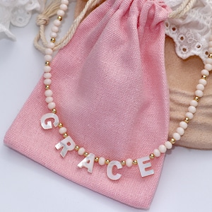 Peut inclure: Un collier de perles composé de petites perles rondes rose pâle alternées avec de minuscules sphères métalliques dorées. Le collier comporte des perles lettres blanches en nacre formant le prénom GRACE, centrées sur le cordon.