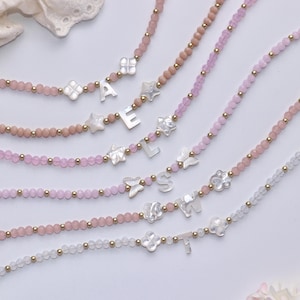 May include: Several beaded necklaces in shades of pink and white, with gold accents. Each necklace features a different letter, along with decorative charms like butterflies, stars, and flowers. The necklaces are displayed on a white surface.