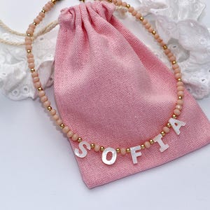 Può includere: Una collana di perline composta da piccole perline opache color pesca, minuscoli distanziatori metallici dorati e perline a lettera in madreperla bianca che compongono il nome SOFIA, appoggiata su un sacchetto di stoffa rosa con bordo in pizzo.