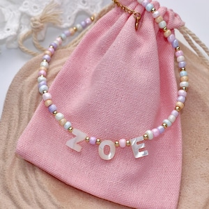 May include: A beaded necklace featuring pastel-colored round beads in shades of pink, blue, and white, interspersed with small gold-toned metallic beads. The necklace includes three white, flat, letter-shaped beads spelling out the name ZOE.