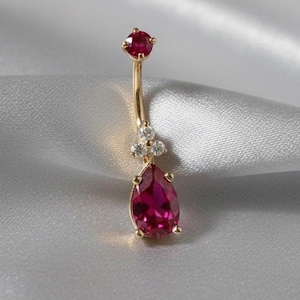 Op de afbeelding: Een gouden navelpiercing met een druppelvormige, dieprode edelsteen. De ring heeft een kleine, ronde rode edelsteen aan de bovenkant en drie kleine heldere edelstenen in het midden. De sieraden zijn geplaatst tegen een zachte, zilverachtige achtergrond.