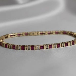Pode incluir: Uma pulseira de ouro com pedras vermelhas retangulares alternadas e pequenas gemas claras e redondas. A pulseira está sobre uma superfície branca. A joia é um estilo de pulseira de tênis.
