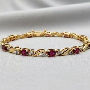 Op de afbeelding: Een gouden armband met afwisselend ovale rode edelstenen en kleine, vierkante, heldere edelstenen. De armband heeft een delicaat, golvend ontwerp en een veilige sluiting. De sieraden worden gepresenteerd op een wit oppervlak.