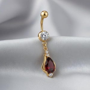 May include: Gold-coloured belly button ring with a curved barbell design. The top ball and a small round gem are clear. A teardrop-shaped, deep red gem hangs from the centre, accented by smaller clear gems.