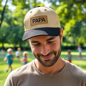 May include: Tan baseball cap with a black brim. The cap features a rectangular patch with the word "PAPA" in bold, black letters. The background shows a green, blurred park setting.