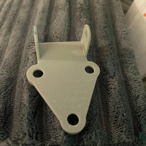 May include: A light gray metal bracket with a triangular base and two vertical extensions. The bracket has three circular holes and two smaller holes at the top. The surface shows minor wear.