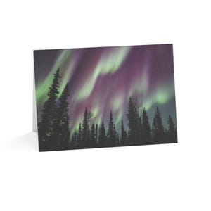 Könnte beinhalten: Grußkarte mit einem lebendigen Bild der Aurora Borealis. Das Bild zeigt eine dunkle Silhouette eines Waldes vor dem Hintergrund wirbelnder grüner und violetter Lichter am Nachthimmel. Die Karte ist weiß.