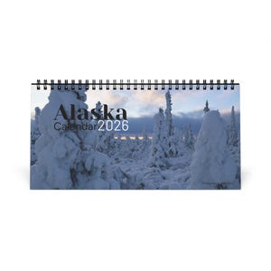 Peut inclure: Calendrier de bureau à spirale avec un paysage enneigé de l'Alaska. La couverture affiche le texte "Alaska Calendar 2026" en noir. L'image montre des arbres enneigés sous un ciel nuageux.